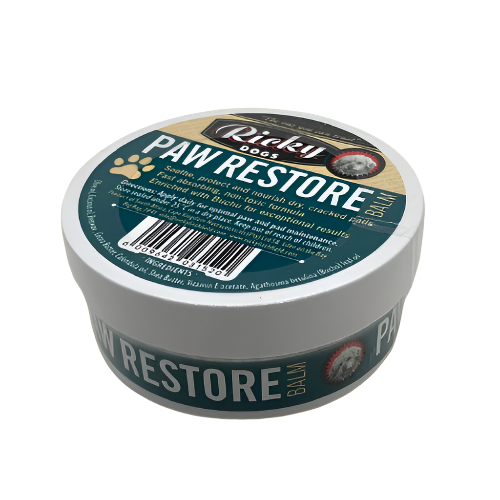 A tub of Paw Restore balm for pets with a label that includes the product name and information such as 'Optimum paw and pad maintenance', 'Heals, soothes and protects rough paw pads', and 'Restores dry/cracked skin'. The label also features a paw print and the brand Ricky Litchfield.